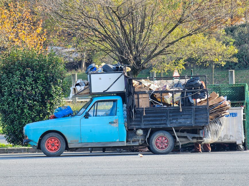 A small, light blue flatbed truck parked on the side of a street, loaded with a large pile of assorted waste materials, including cardboard boxes, black trash bags, and various other debris. The truck's bed is surrounded by a black metal railing, with some of the waste secured by ropes. The vehicle’s wheels are red, and the front of the truck shows signs of wear and rust, with the vehicle positioned parallel to a curb. Behind the truck, a green bush partially obscures the left side, and a large tree with a full canopy of leaves is visible in the background, suggesting a suburban or park area. The scene is illuminated by natural daylight, and the setting hints at a private or independent waste removal service, such as those offered by Rubbish Clearance Merton, facilitating on-site clearance and alternative waste handling outside of local authority rubbish collection. The overall environment appears calm, with no active loading or unloading visible.