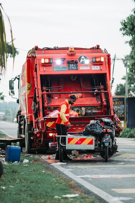 A worker wearing a high-visibility yellow and red uniform is operating a red rubbish collection vehicle parked on the side of a street, with the rear loading compartment open. The worker is leaning forward, handling debris or waste that has been collected from the pavement area. Nearby, there is a small black motorcycle or scooter parked on the sidewalk with a black plastic bag resting on its seat, likely filled with waste. The scene takes place in an urban environment with a sidewalk, street lane markings, and some greenery including trees and grass visible in the background. The rubbish truck, labeled with service markings and safety indicators, appears to be in the process of an independent or private waste collection, reflecting how companies like Rubbish Clearance Merton offer on-site clearance and alternative waste handling services. The overall atmosphere is typical of a busy street scene focused on rubbish removal operations, with natural daylight illuminating the scene and some distant structures or signage also visible in the background.