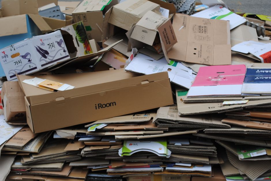 A large pile of discarded cardboard and paper packaging materials, including flattened boxes, unassembled boxes, and crumpled sheets, is stacked outdoors on a paved surface. The cardboard varies in color from light brown to darker shades and includes some printed and branded packaging, such as a box labeled 'iRoom.' The materials appear weathered, with some flattened but others partially folded or folded over, indicating recent collection or sorting. Surrounding the main heap are additional loose cardboard pieces and paper waste, with some items overlapping and spilling onto the ground. The environment appears to be a waste collection or disposal site, reflecting the type of materials handled by independent rubbish removal services, such as Rubbish Clearance Merton, for business or household waste management. The scene is well-lit with natural lighting, emphasizing the textures and condition of the packaging materials, highlighting the needs for professional waste clearance and proper disposal.