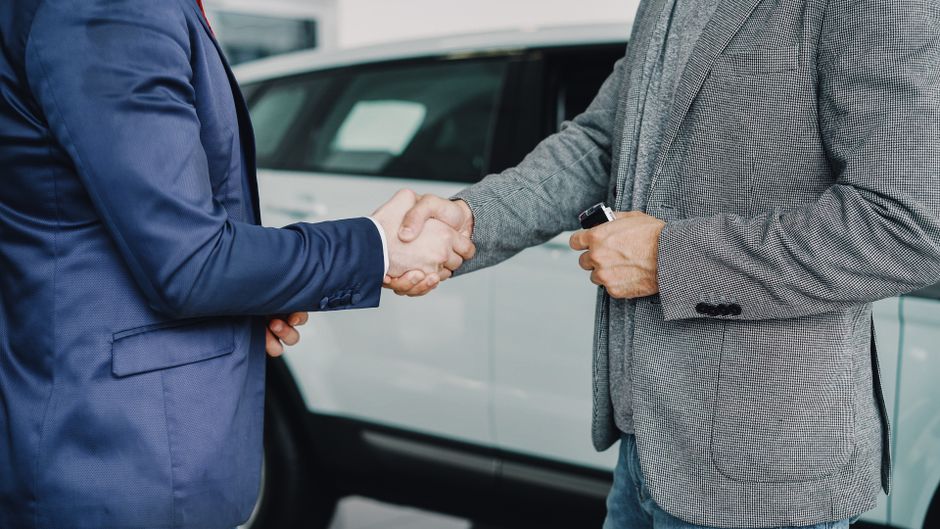 A close-up image showing two men engaged in a handshake outdoors, with one dressed in a navy blue suit and the other in a grey checked blazer. The man in the suit has his left hand resting on his side, while the man in the blazer holds a set of keys or key fob in his right hand. They are standing next to a white vehicle parked on a paved surface, possibly a driveway or street, with the body of the vehicle visible in the background. The scene is well-lit, indicating daylight, and the environment suggests a professional or business setting, such as a commercial or waste management context, where private arrangements for rubbish removal or waste disposal may be discussed, possibly involving services like those provided by Rubbish Clearance Merton for local businesses like those in Wimbledon.