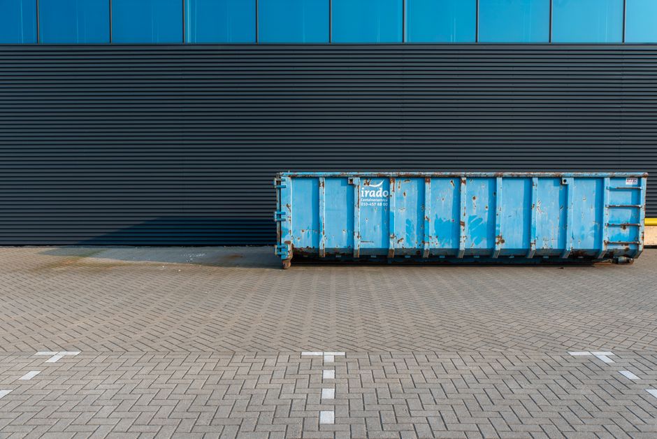 A large, rectangular blue metal skip with visible rust and chipped paint sits on a paved outdoor area, positioned against a modern building with dark horizontal siding and blue-tinted glass panels. The skip's surface shows signs of wear, including rust spots and scratches, and is situated near a yellow safety barrier on its right. The surrounding paving consists of small, interlocking concrete bricks arranged in a linear pattern, with marked parking spaces visible in the foreground. The background environment appears industrial or commercial, providing an unobstructed view of the skip's purpose for waste storage or rubbish removal, consistent with services offered by Rubbish Clearance Merton in managing commercial waste contracts for local businesses.