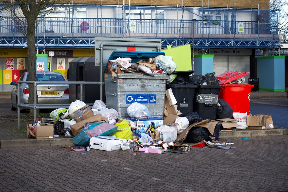 An overflowing waste disposal area in an urban setting, featuring a large grey mixed paper and cardboard recycling bin positioned on a paved sidewalk, with its lid open and various paper products, cardboard boxes, and plastic bags spilling onto the ground in front. To the left of the bin, there are smaller black rubbish bags and additional cardboard boxes, some flattened and others partially collapsed. A red waste bin is situated nearby, also containing waste materials. In the background, there is a street with parked cars, including a silver vehicle with a visible license plate, and a building with a blue scaffolding structure and a shopfront displaying various signs. The scene is illuminated by natural daylight, emphasizing the collection of mixed waste and packaging materials, indicative of a private waste collection or rubbish removal service often seen in urban environments. Rubbish Clearance Merton provides such services for local clearance and waste disposal needs in the area.