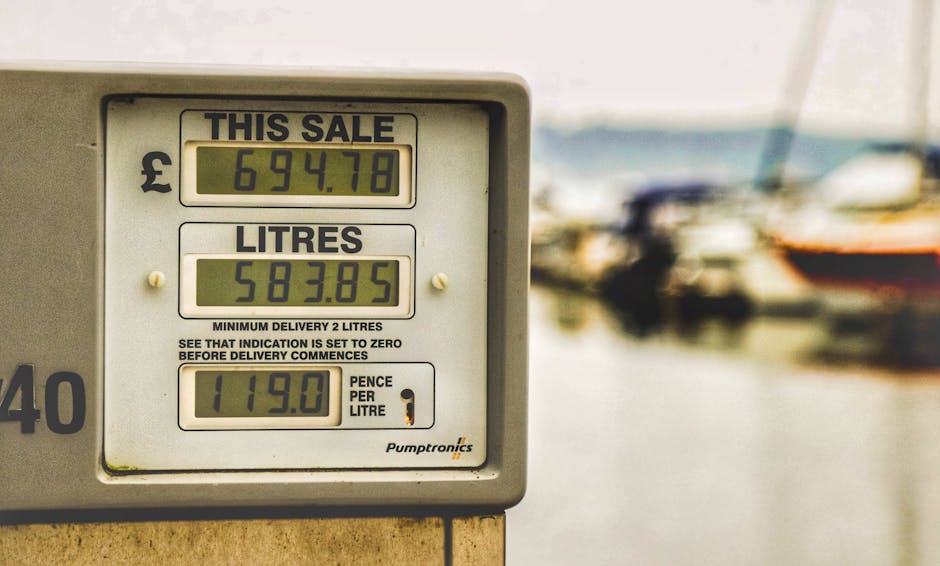 Close-up view of a digital fuel meter displaying the total cost of £694.78 for a full tank, with the amount of fuel currently in the tank shown as 583.85 litres and a fuel price of 119.0 pence per litre, mounted on a beige or light-colored panel. The background is slightly blurred, revealing a marina or boatyard environment with several boats docked on the water, some with visible dark hulls and cabins, and a cloudy sky above. The scene is illuminated by natural daylight, emphasizing the utilitarian appearance of the fuel meter, which may be associated with private vessel refuelling or on-site fuel management for boats. This image connects to topics of independent fuel and waste management, relevant for professional rubbish removal services like Rubbish Clearance Merton, highlighting the practical aspects of responsible disposal and resource handling outside of standard municipal waste channels.