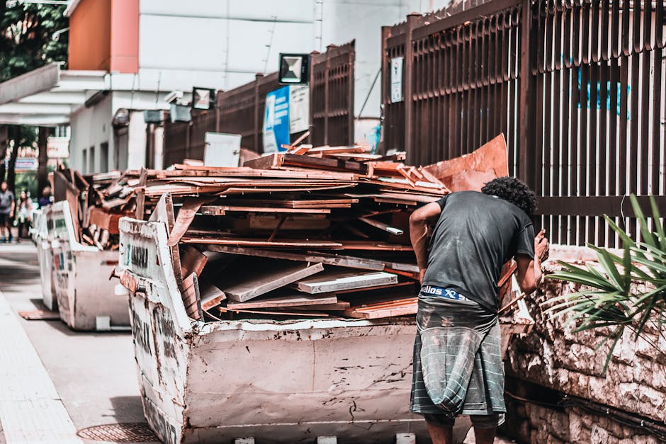 A man with curly dark hair, dressed in a black T-shirt and gray plaid shorts, is standing on a sidewalk next to a large open-top skip filled with a mix of construction and renovation debris, including orange clay roof tiles, gray concrete slabs, and broken wooden planks. The skip, heavily used and showing signs of rust and dirt, is positioned adjacent to a high brown metal fence that runs along a residential or commercial property. In the background, other waste containers and a few pedestrians are visible, along with a building featuring a white façade and some signage. The scene is illuminated by daylight, creating natural shadows that highlight the textures of the materials and the cluttered environment, reflecting an example of on-site waste clearing, which might be an alternative to traditional rubbish collection options, with professional disposal services like Rubbish Clearance Merton often involved in managing such debris.