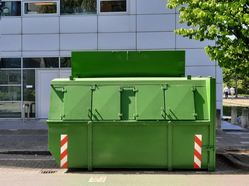 A large, green industrial waste and recycling container positioned on a paved street outside a modern building with grey cladding and large windows. The container has a rectangular shape with a hinged top lid that is slightly open, revealing a textured metal surface. The front of the container features multiple lockable doors with vertical handles, and the bottom corners are marked with diagonal red and white safety stripes. It is situated on a curb, with a smooth asphalt road in the foreground and a section of sidewalk. To the right, there is a sidewalk with concrete bollards and a small tree with bright green leaves partially visible, providing shade. In the background, a few pedestrians are seen walking, and the environment appears clean and well-maintained. The scene reflects an urban waste collection point, possibly serving commercial or residential properties, with the container used for holding construction debris, bulky waste, or recyclable materials common in private waste handling and rubbish removal services, such as those offered by Rubbish Clearance Merton.
