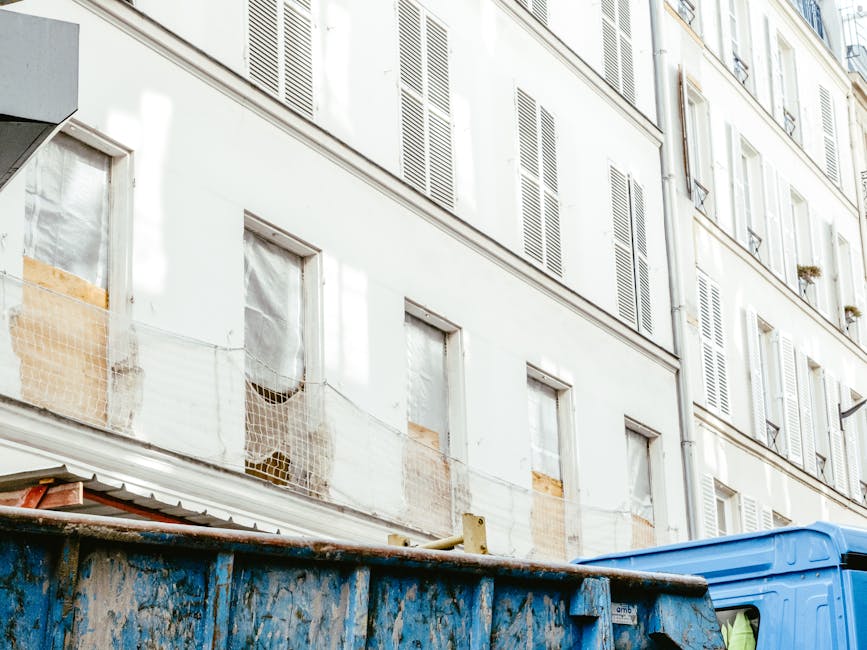 A man with curly dark hair, dressed in a black T-shirt and gray plaid shorts, is standing on a sidewalk next to a large open-top skip filled with a mix of construction and renovation debris, including orange clay roof tiles, gray concrete slabs, and broken wooden planks. The skip, heavily used and showing signs of rust and dirt, is positioned adjacent to a high brown metal fence that runs along a residential or commercial property. In the background, other waste containers and a few pedestrians are visible, along with a building featuring a white façade and some signage. The scene is illuminated by daylight, creating natural shadows that highlight the textures of the materials and the cluttered environment, reflecting an example of on-site waste clearing, which might be an alternative to traditional rubbish collection options, with professional disposal services like Rubbish Clearance Merton often involved in managing such debris.