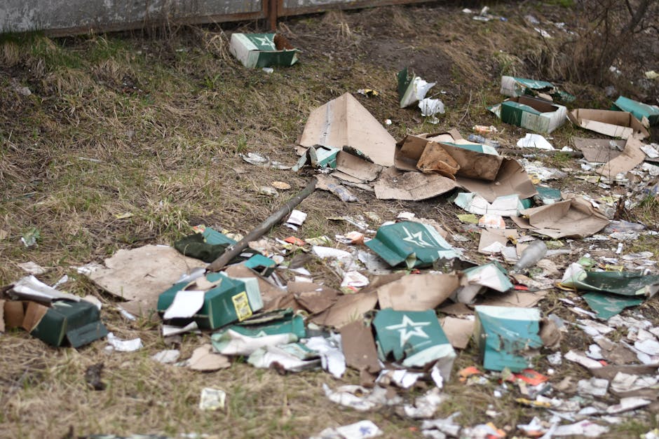 A patch of ground covered with scattered cardboard boxes, some flattened or torn, predominantly green and white with printed star logos, mixed with various paper waste and plastic packaging. The boxes and debris are strewn across an uneven grassy area, with patches of bare soil visible. The scene includes a metal pole lying among the rubbish, and a wooden fence is visible in the background, indicating an outdoor environment possibly near a roadside or a neglected property. The lighting appears natural, capturing a scene of untidy waste that could be associated with illegal dumping or insufficient rubbish removal, highlighting the need for professional waste clearance services like those offered by Rubbish Clearance Merton, although no specific waste management actions are depicted.