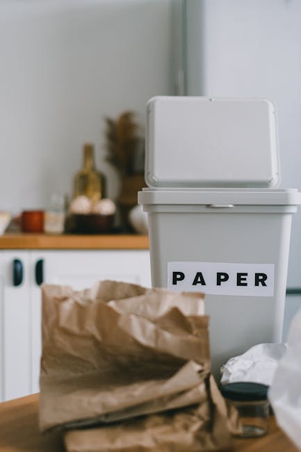 A tall, rectangular white paper recycling bin with a partially open lid, situated on a wooden surface in a domestic setting. The bin has a clear label reading 'PAPER' in bold black letters affixed to its front. In front of the bin, there is a crumpled brown paper bag and some loose white paper or tissue, indicating household waste that is ready for collection or disposal. The background features a modern kitchen environment with light-colored cabinetry, including visible black handles, and a blurred view of kitchen items such as bottles and jars on a countertop, suggesting an indoor space dedicated to household rubbish management, reflecting a typical example of informal waste separation at home, aligned with curbside or private waste collection practices managed by companies like Rubbish Clearance Merton.