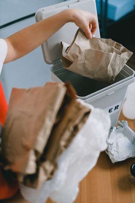 A person is shown placing a brown paper bag into a small, grey, open-top rubbish bin with a black interior, located on a wooden floor. The individual’s arm is visible, extending from the left, and they are wearing a short-sleeved shirt. In the foreground, several crumpled paper bags are held in the person's other hand. The background is slightly blurred, with a focus on the individual and the rubbish bin, which is labeled with a partially visible word, suggesting a designated waste container. The scene appears to be in an indoor environment, such as a kitchen or storage area, emphasizing the process of managing household waste or preparing for rubbish collection. The natural lighting highlights the textures and colors of the paper waste, the bin, and the person's skin, with minimal distractions, conveying a clear view of rubbish disposal activity relevant to private waste handling and rubbish removal services like those provided by Rubbish Clearance Merton depicted naturally within an everyday context.