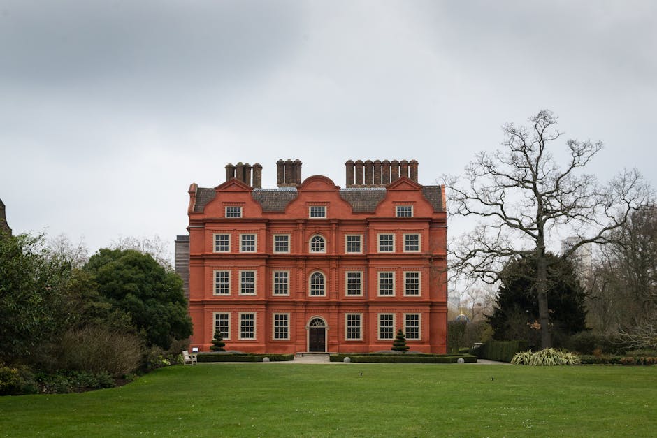A large, four-story red brick building with a symmetrical facade situated in a landscaped garden area. The building features multiple white-framed sash windows on each floor, a central arched doorway, and decorative brickwork beneath the windows. The roof is adorned with several chimney stacks, and the gabled roof sections are finished with grey slate tiles. In the foreground, there is a well-maintained lawn with neatly trimmed grass, and two small conical shrubs flank the entrance path. To the right of the building, a tall, leafless tree extends its bare branches into the overcast sky, while on the left, dense green bushes partially obscure the view of the side of the structure. The environment suggests a formal, historical residential or institutional property, with natural surroundings including other trees and garden elements, creating an overall scene consistent with private estate grounds potentially requiring waste management or clearance services. Rubbish Clearance Merton sometimes undertakes on-site clearance or private rubbish removal tasks in similar landscaped settings, emphasizing the importance of sustainable disposal of accumulated waste or debris.