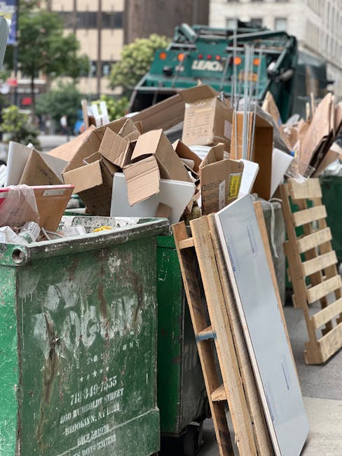 An overflowing green rubbish skip filled with various types of waste including cardboard boxes, some flattened and others in their original shape, along with several pieces of wood, including a partially assembled wooden pallet leaning against the side of the skip. The cardboard boxes are of different sizes, with some standing upright and others toppled, showing their corrugated texture and brown color. The wood appears unfinished, with visible grain and rough edges. In the background, a recycling truck with the logo that is partly visible is parked on a city street surrounded by urban buildings, trees, and parked cars, suggesting a typical setting for waste collection or on-site clearance. The scene is lit naturally, presenting a clear, daytime environment indicative of private waste handling as part of an independent rubbish removal service by Rubbish Clearance Merton, capturing the practical aspect of rubbish disposal in urban areas.