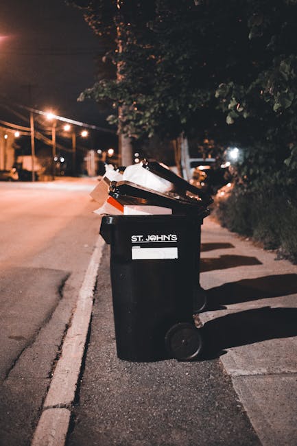 A black plastic rubbish bin with the label 'St. John's' positioned on a paved sidewalk next to a curb in an urban street setting during evening hours. The bin is filled with various waste materials, including cardboard, paper, and plastic items, some of which are partially visible and protruding from the open lid. The surrounding environment features street lighting casting warm glow, with a background of trees, roadside vegetation, and faint streetlights, indicating a residential or mixed-use area. The scene suggests an example of private or independent waste disposal, aligning with rubbish removal services such as those offered by Rubbish Clearance Merton for local waste clearance needs. The texture of the bin’s surface appears smooth and slightly reflective, while the waste inside shows crumpled paper and flattened cardboard with varied textures, contrasting with the rough asphalt and sidewalk surface. Overall, this image highlights the concept of on-site rubbish collection and private waste management outside typical municipal schemes, emphasizing the importance of proper disposal through commercial rubbish removal services in urban environments.