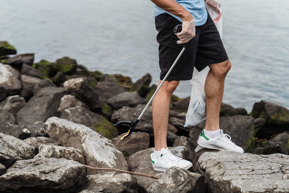 The image shows a person standing on a rocky shoreline near a body of water, with only the lower half of their body visible. They are wearing black shorts, white sports shoes with green accents, and a light blue t-shirt. The individual is holding a long-handled grabber tool in their right hand and appears to be in the process of collecting litter or debris from the rocks. A white plastic bag is hanging from their left hand, suggesting that they are engaged in a waste collection activity. The rocks are irregular in shape, with varying sizes and textures, some covered with patches of green moss or algae, indicating the damp environment typical of a seaside or riverbank. The background shows the water's surface, slightly rippled, with an overcast sky casting diffused light over the scene. This setting reflects an outdoor effort in waste management or environmental cleanup, consistent with services offered by companies like Rubbish Clearance Merton for private or alternative waste disposal, emphasizing on-site rubbish collection and environmental responsibility.