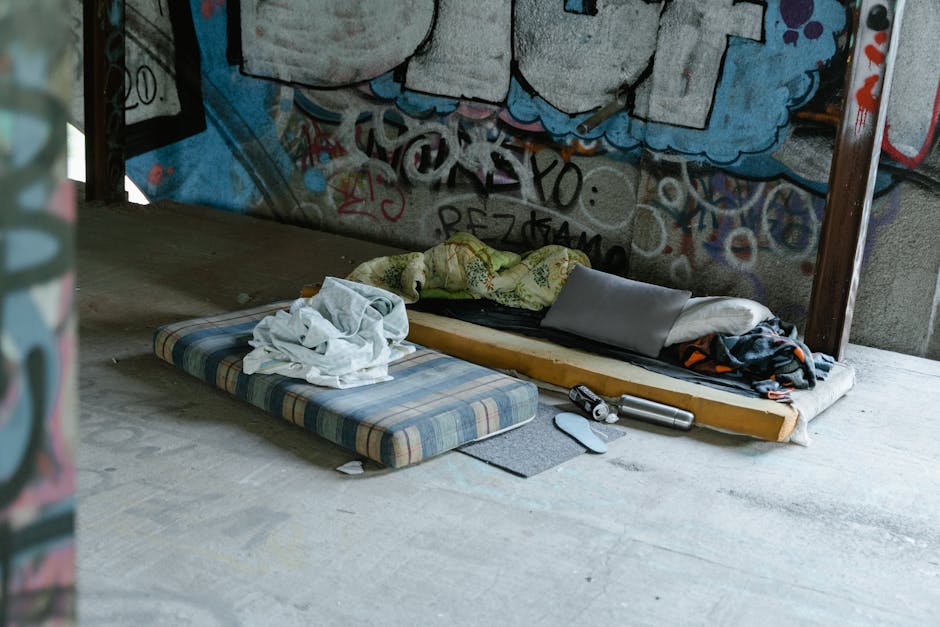 In a dimly lit indoor setting with graffiti-covered walls, two makeshift bedding setups are visible on a concrete floor. The first consists of a thin, striped foam mattress with a crumpled white bedsheet placed on top. Nearby, there is a small gray foldable cushion or pillow. The second bedding is a worn-out, mattress with a wooden frame underneath, covered partially by a light-colored blanket and a dark-patterned cloth. A single pillow rests on top of this mattress. Scattered around the bedding, there is a stainless steel thermos, a set of keys, and a small gray item, possibly a shoe or footwear. The scene appears to be a casual or temporary resting place, likely in an abandoned or underused building, emphasizing a context where independent rubbish removal or on-site clearance might be necessary to manage clutter and waste associated with such environments. Rubbish Clearance Merton offers services suited for cleaning and waste disposal in these types of settings, supporting private or alternative waste handling solutions.