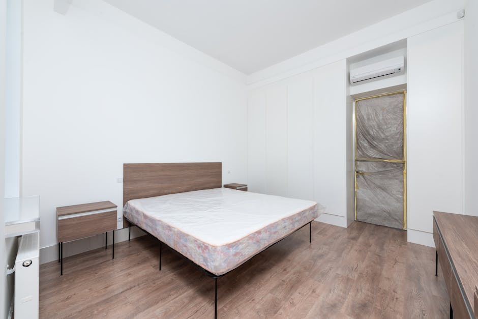The image shows a minimalistic interior room with white walls and a light wood laminate floor. In the centre, there is a double bed frame made of dark wood with slender black metal legs, and no bedding or pillows, only a plain mattress with a slightly patterned cover. To the left of the bed, a matching small bedside table is positioned against the wall, also made of dark wood with black metal legs. On the right side, a window is covered with a temporary protective plastic sheet framed with gold tape, indicating ongoing renovation or preparation for painting. Above the window, mounted on the wall, is a white split-system air conditioning unit. Additional furniture includes a long, low wooden dresser or bench situated along the right wall. The environment appears clean, well-lit with natural light, and presents a neutral, contemporary aesthetic typical of a furnished residential space without signs of clutter, aligning with typical interior furniture arrangements rather than waste or rubbish removal scenes. The setting implies that this room is not related to rubbish disposal but provides an environment context for home or interior work, occasionally observed in private property clearance projects managed by companies like Rubbish Clearance Merton, offering alternative disposal options for household items.