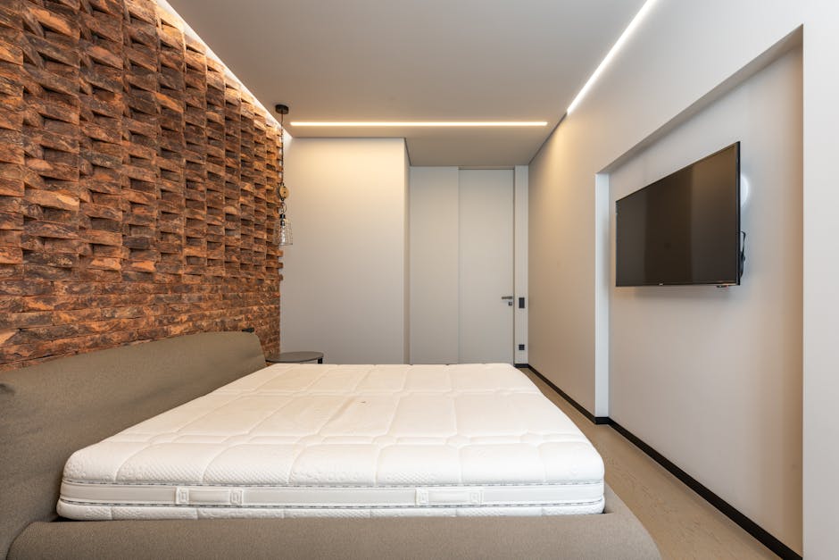 In a dimly lit indoor setting with graffiti-covered walls, two makeshift bedding setups are visible on a concrete floor. The first consists of a thin, striped foam mattress with a crumpled white bedsheet placed on top. Nearby, there is a small gray foldable cushion or pillow. The second bedding is a worn-out, mattress with a wooden frame underneath, covered partially by a light-colored blanket and a dark-patterned cloth. A single pillow rests on top of this mattress. Scattered around the bedding, there is a stainless steel thermos, a set of keys, and a small gray item, possibly a shoe or footwear. The scene appears to be a casual or temporary resting place, likely in an abandoned or underused building, emphasizing a context where independent rubbish removal or on-site clearance might be necessary to manage clutter and waste associated with such environments. Rubbish Clearance Merton offers services suited for cleaning and waste disposal in these types of settings, supporting private or alternative waste handling solutions.