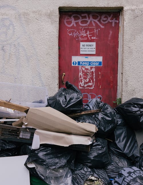 A collection of black garbage bags and assorted discarded waste materials piled in front of a red metal door, which features graffiti tags, a 'No Money' notice, and a 'Keep Clear' sign. The bags and debris include cardboard boxes, foam packaging, and other refuse, situated on a pavement next to a beige, textured building wall. The scene indicates an outdoor rubbish accumulation, potentially related to private clearance or local waste handling, with the waste temporarily stored outside a building entrance. The lighting appears natural, highlighting the clutter and the urban setting around the property. This image could be associated with independent rubbish collection services, such as those offered by Rubbish Clearance Merton, especially in the context of resolving fly-tipping issues in residential or commercial streets.