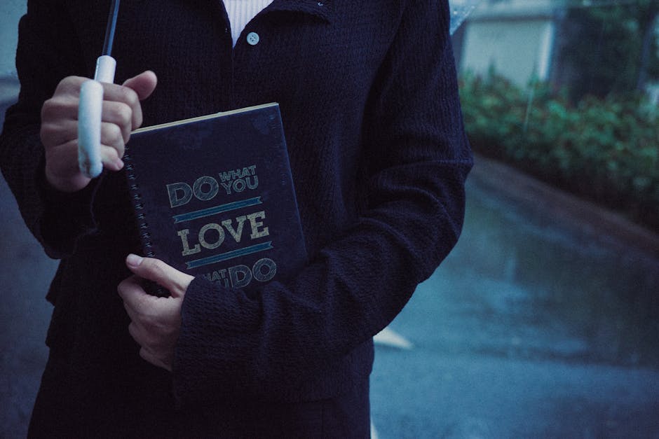 A person dressed in a dark navy sweater and black jacket is holding a small, dark-colored spiral-bound notebook with the phrase 'Do What You Love' printed in bold, light-colored letters on the cover. The individual is standing outdoors on a paved surface, with a blurred background that includes greenery and a building with windows, suggesting a residential or suburban street. The person's right hand grips the notebook, while their left hand holds a white marker or pen, positioned near the top of the notebook. The lighting indicates it is either early morning or late evening, with subdued natural light creating a calm, muted atmosphere. While the main focus of the image is on the person and the notebook, the environment hints at an everyday setting, possibly involving outdoor activities or personal reflection. Although the scene does not directly relate to rubbish removal or waste management, it reflects a casual moment in an urban or suburban context, aligning with themes of everyday life and personal choice.