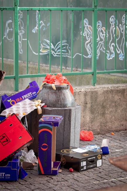 A collection of black garbage bags and assorted discarded waste materials piled in front of a red metal door, which features graffiti tags, a 'No Money' notice, and a 'Keep Clear' sign. The bags and debris include cardboard boxes, foam packaging, and other refuse, situated on a pavement next to a beige, textured building wall. The scene indicates an outdoor rubbish accumulation, potentially related to private clearance or local waste handling, with the waste temporarily stored outside a building entrance. The lighting appears natural, highlighting the clutter and the urban setting around the property. This image could be associated with independent rubbish collection services, such as those offered by Rubbish Clearance Merton, especially in the context of resolving fly-tipping issues in residential or commercial streets.
